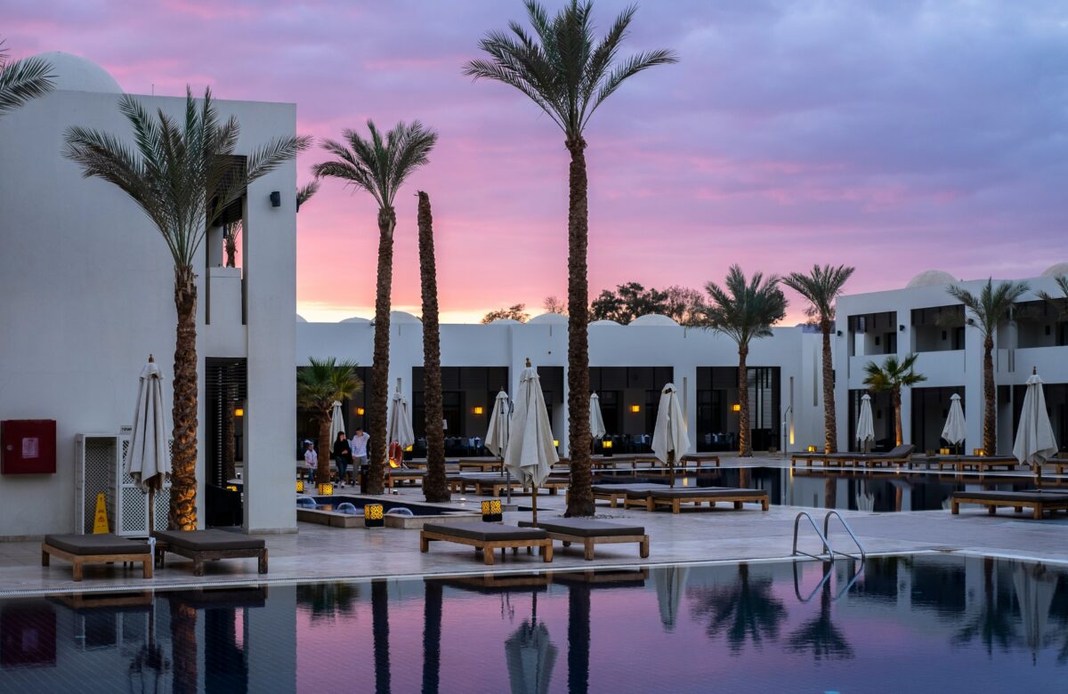 Best hotel credit cards can unlock access to luxury resort pools like this one, with modern white buildings, palm trees, and loungers reflecting in still water under a pink sunset sky